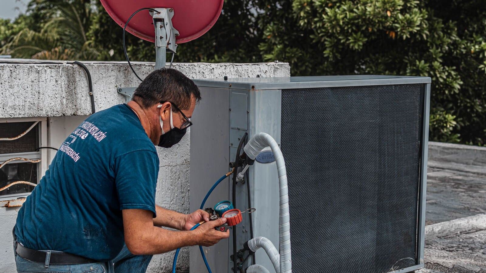 An HVAC technician kneeling on a rooftop, both hands on a manifold gauge while servicing an outdoor air conditioning unit.